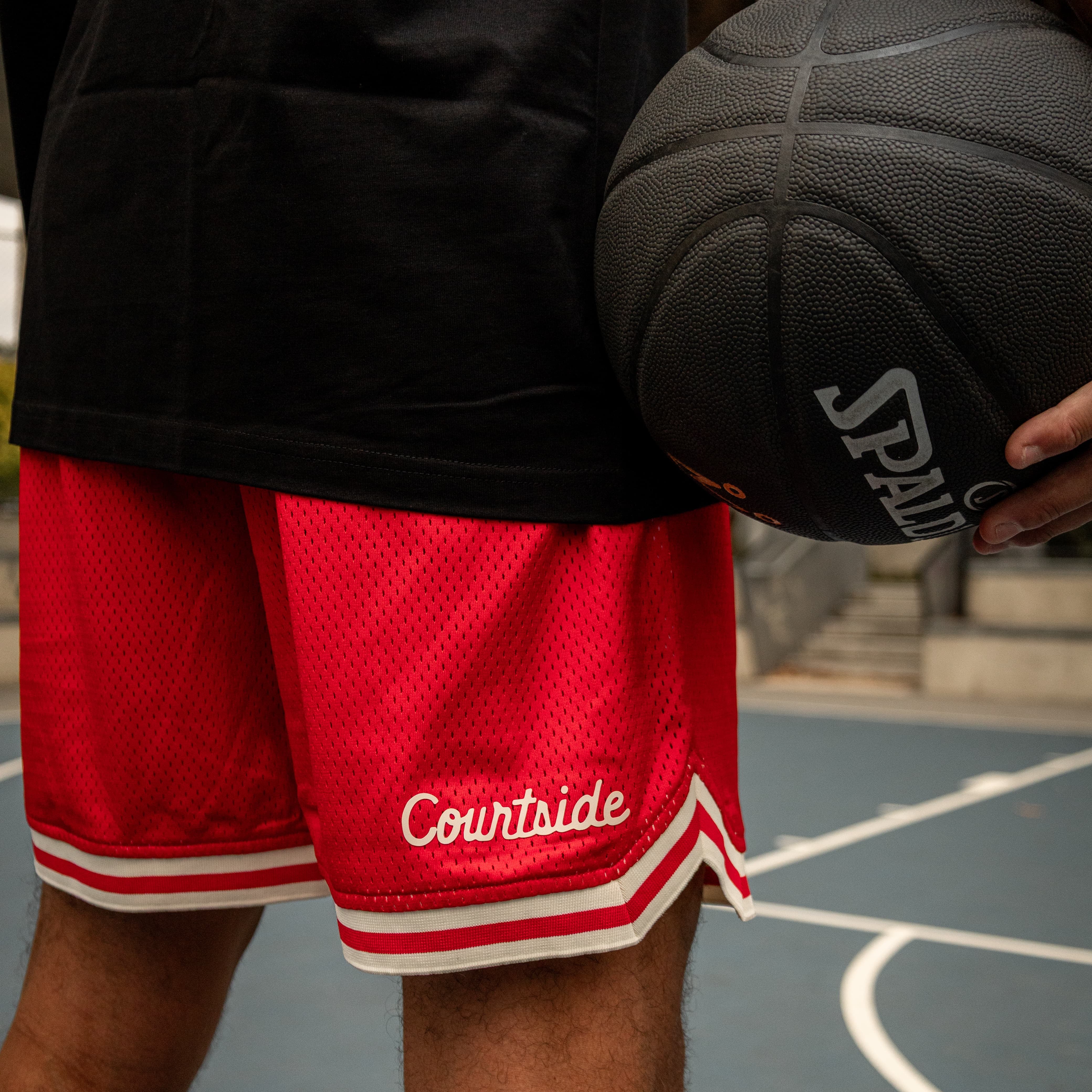 Person wearing red Courtside basketball shorts holding a black basketball on a court.
