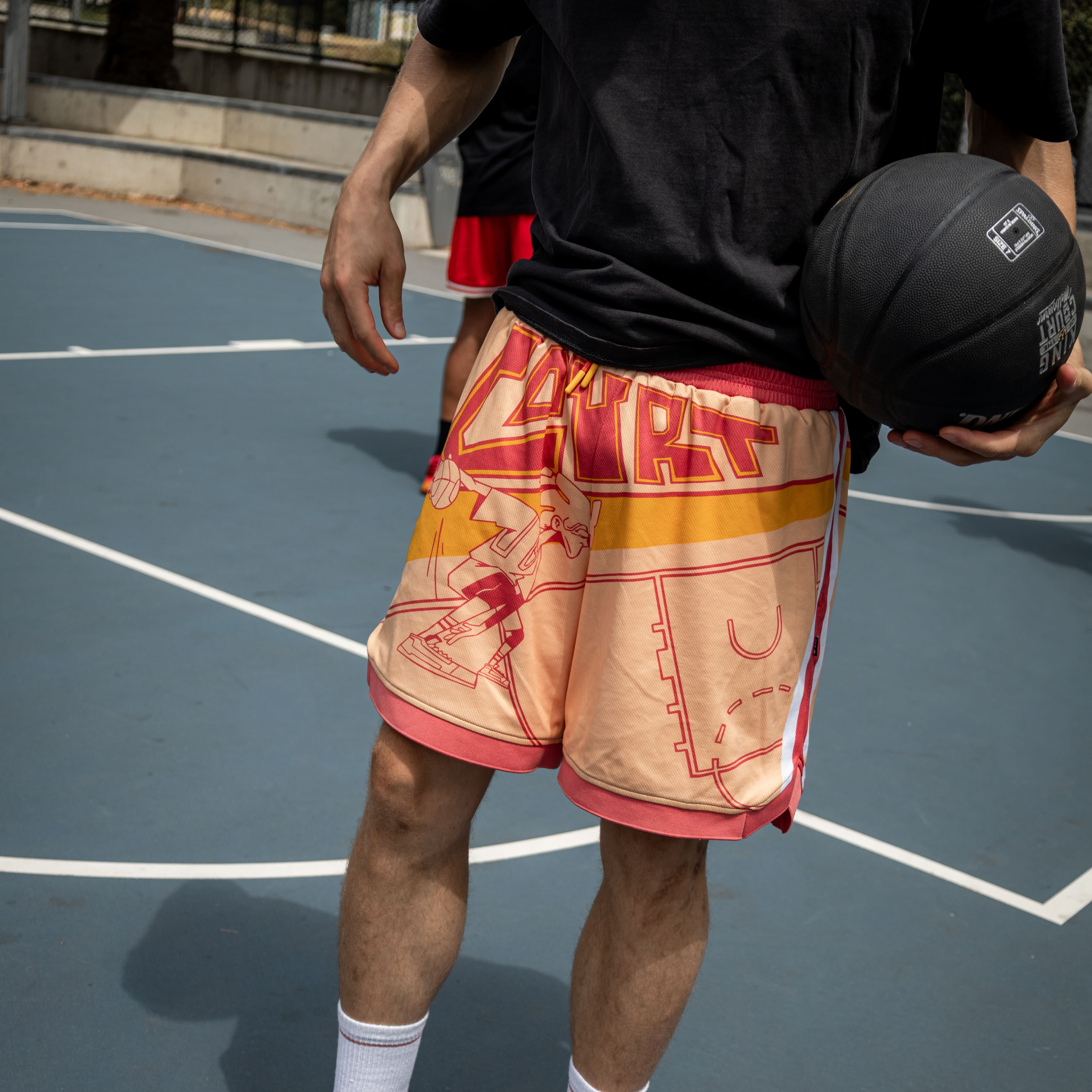 Person holding a basketball on an outdoor court wearing a black Courtside shirt and special edition graphic colourful Courtside shorts.