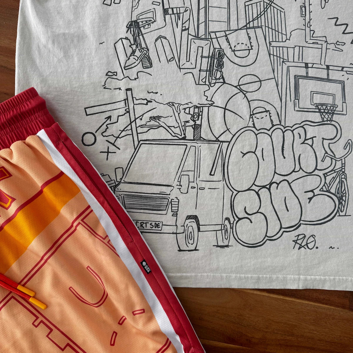 Bone t-shirt with black cityscape graphic and orange shorts on a wooden surface