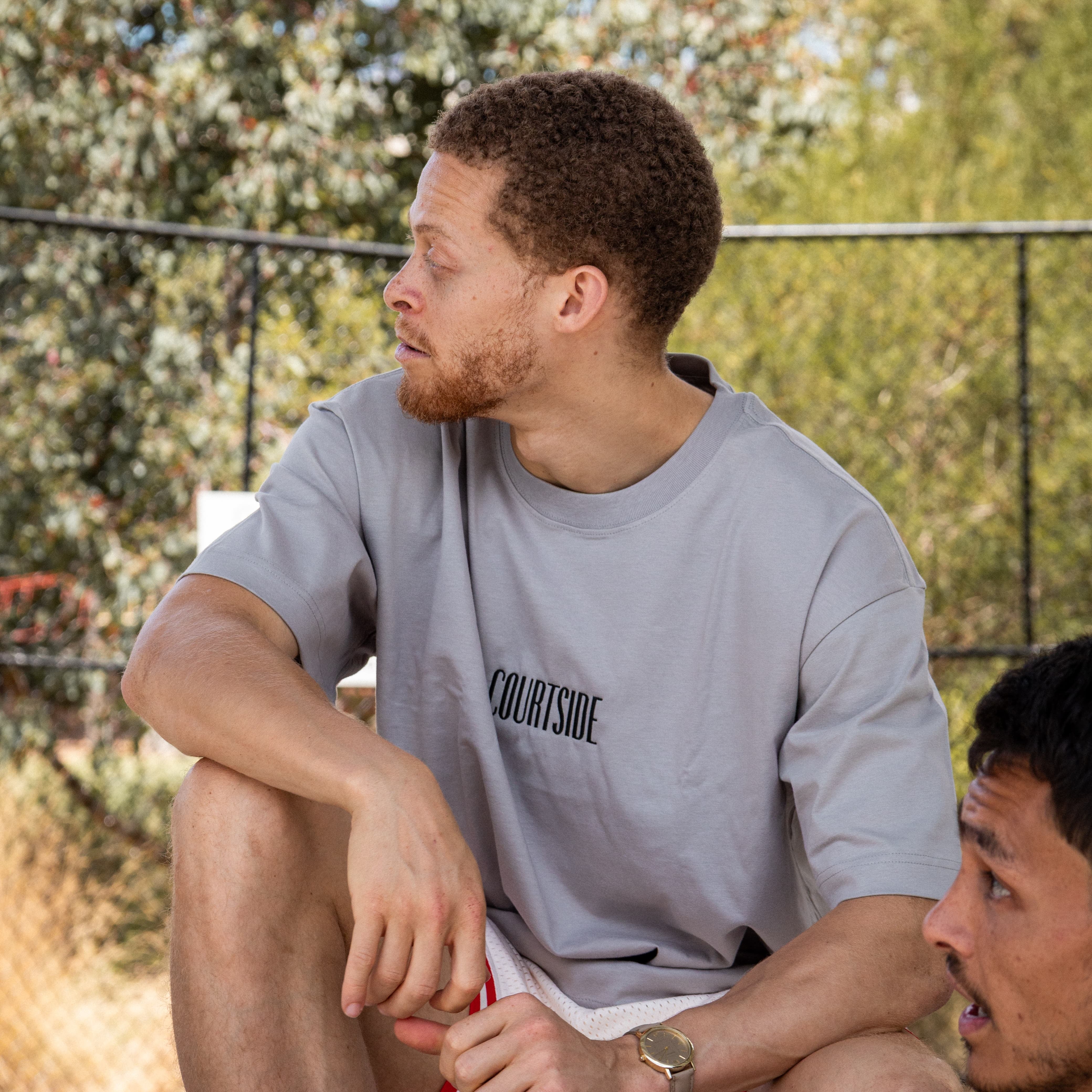 Two men sitting outdoors courtside with trees in the background