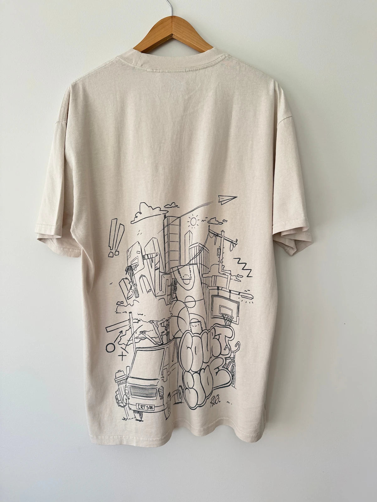 Bone t-shirt with black handdrawn graphic design on back, placed on a wooden hanger against a white wall