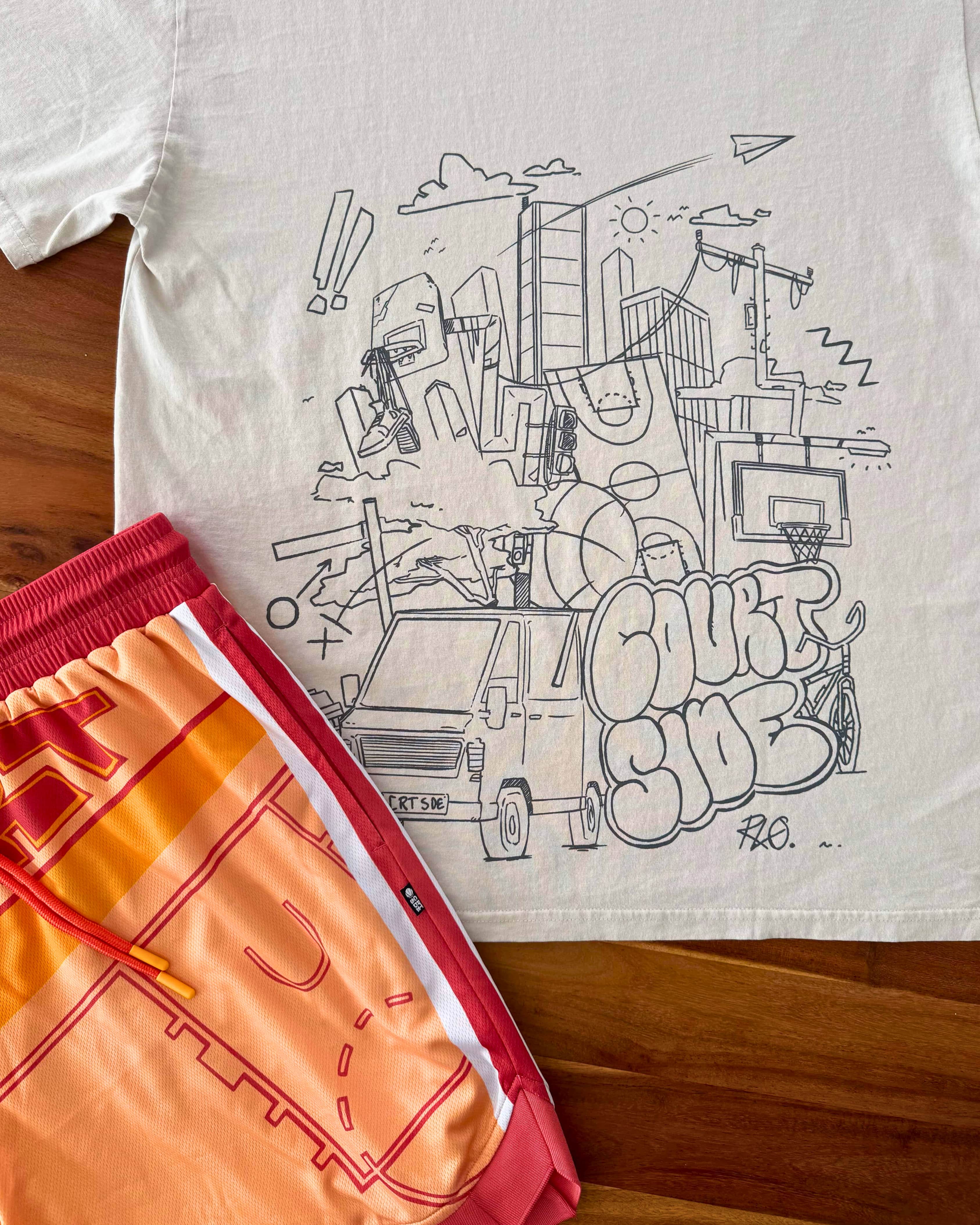 Bone t-shirt with black cityscape graphic and orange basketball shorts on a wooden surface