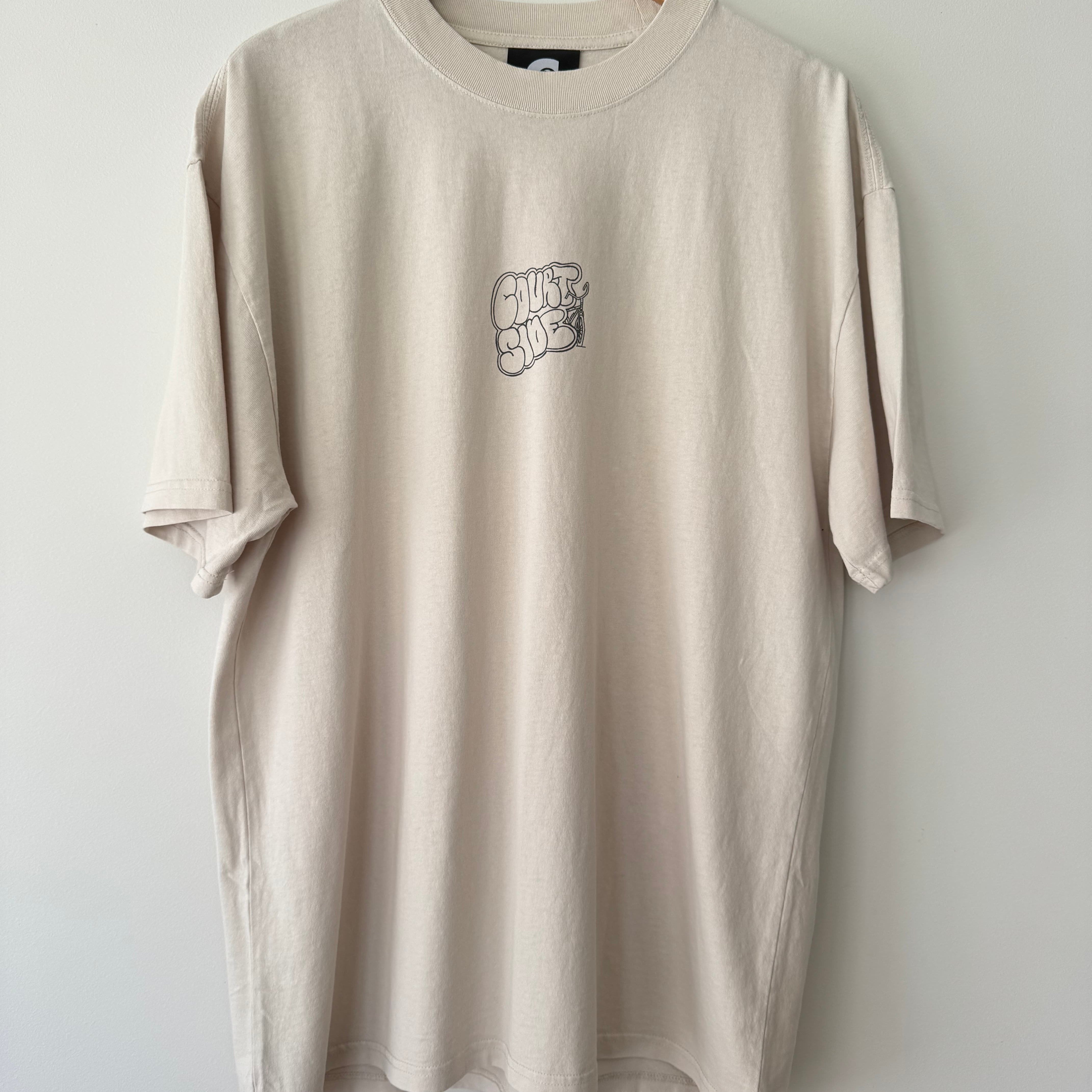 Bone beige t-shirt with a Courtside logo on front chest hanging on a wooden hanger against a white wall