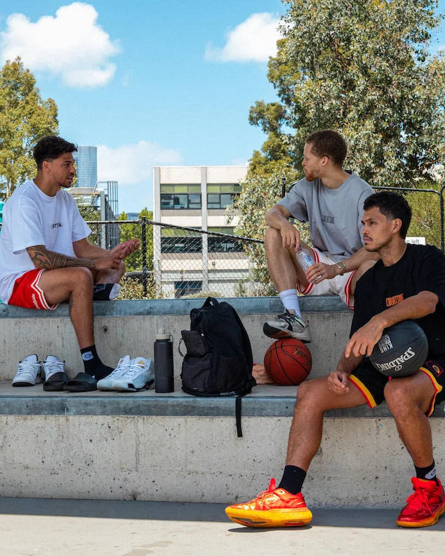 Three basketballers sitting courtside on a concrete ledge with basketball chatting with a clear blue sky and trees in the background.