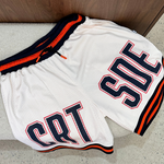 Cream basketball shorts with orange and black accents on a concrete floor.