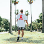 A person walking away from the camera wearing a white t-shirt with a red and logo on the back, in a park setting with palm trees.