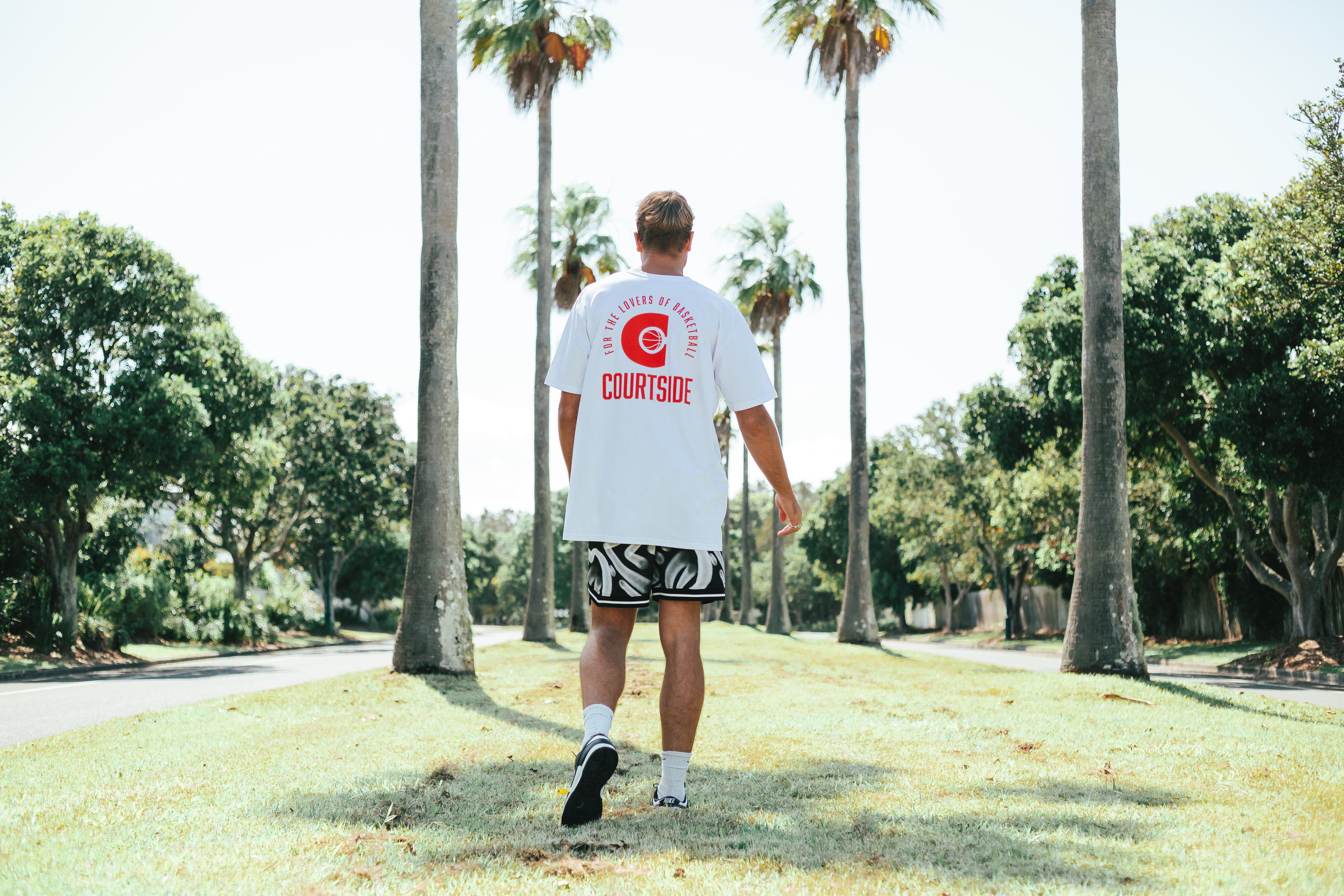 A person walking away from the camera wearing a white t-shirt with a red and logo on the back, in a park setting with palm trees.