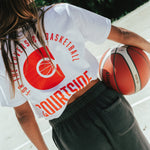 Female wearing a white t-shirt with red detail holding a basketball with her back to camera on an outdoor basketball court