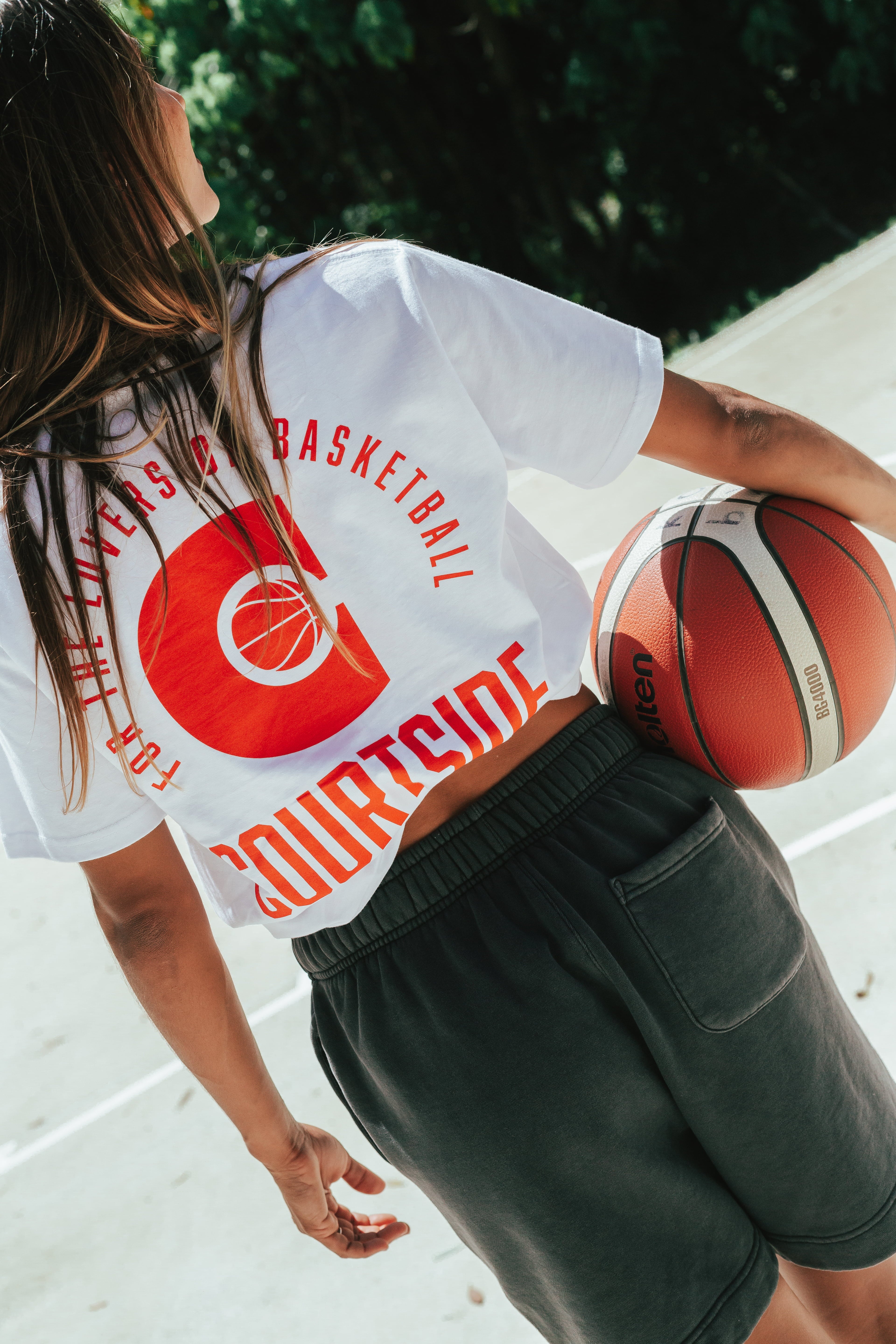 Female wearing a white t-shirt with red detail holding a basketball with her back to camera on an outdoor basketball court
