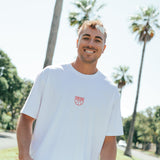 Man smiling wearing a white t-shirt with red centre chest logo with palm tree in the background