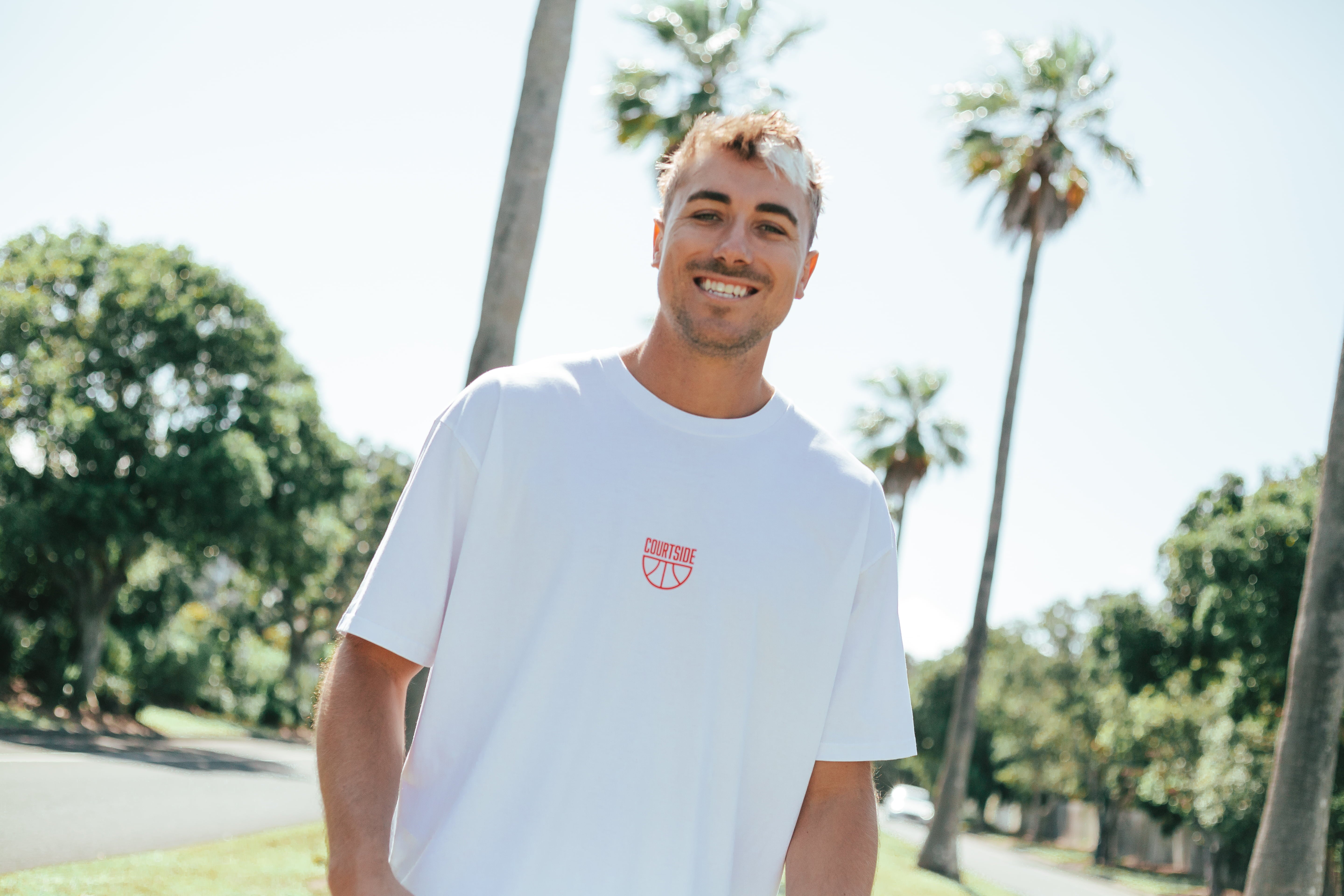 Man smiling wearing a white t-shirt with red centre chest logo with palm tree in the background