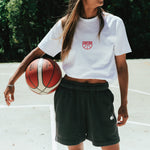 female wearing white tshirt with red centre logo and shorts holding a basketball standing on an outdoor basketball court