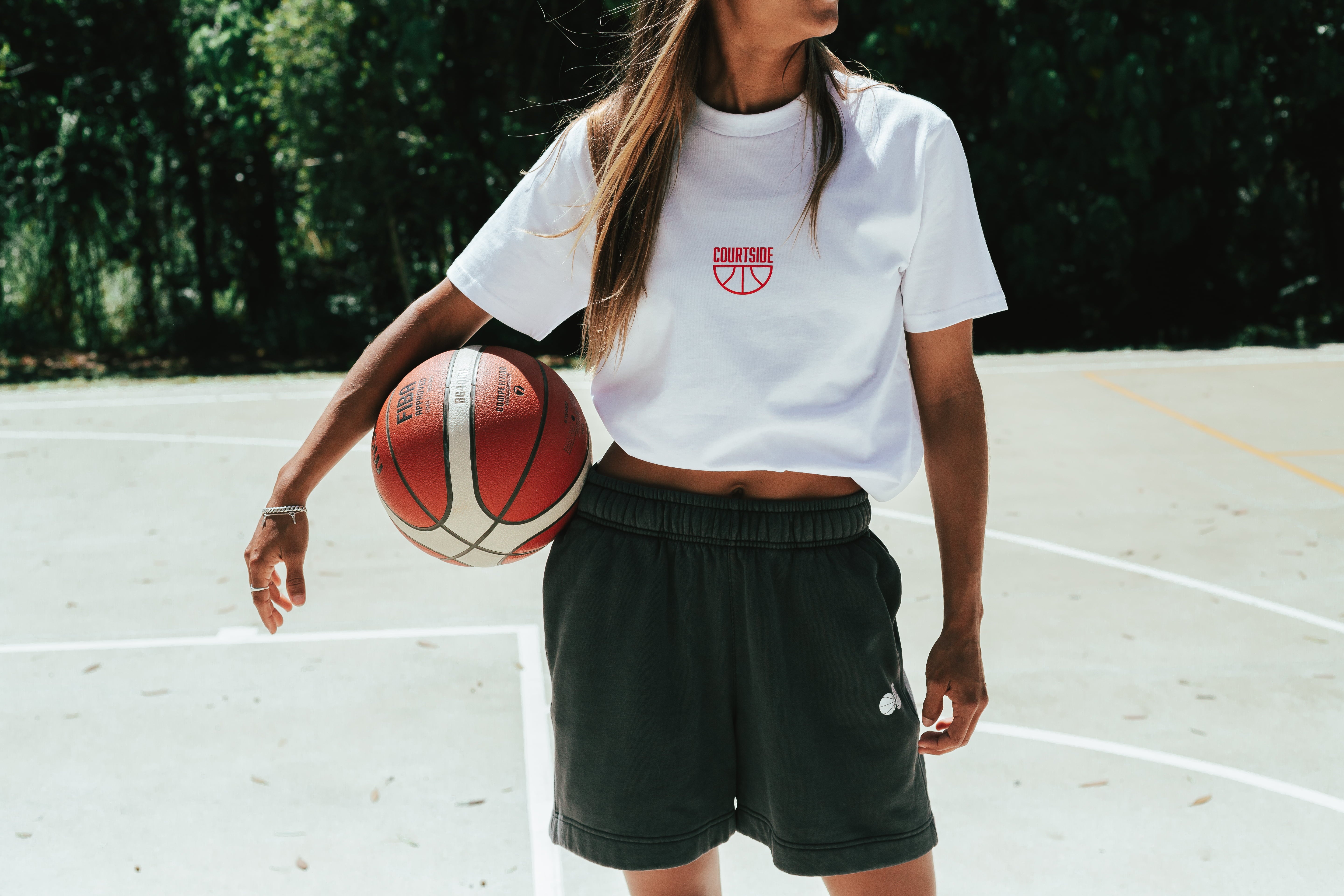 female wearing white tshirt with red centre logo and shorts holding a basketball standing on an outdoor basketball court