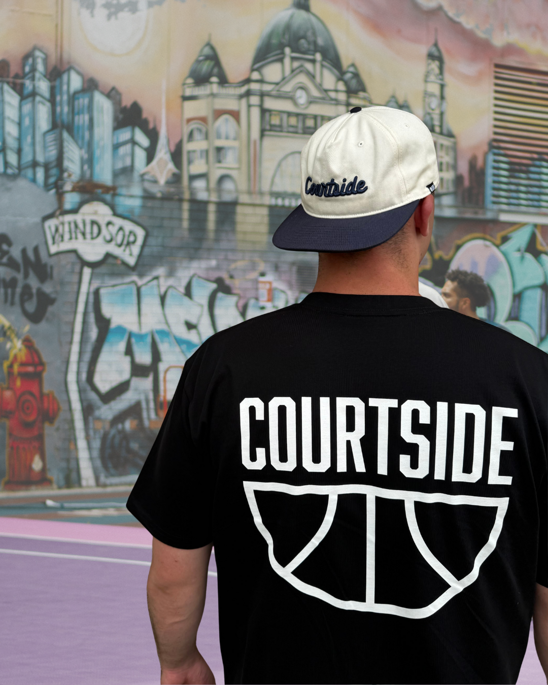 Person wearing a black 'Courtside' t-shirt with a Courtside cap, standing in front of a colourful basketball court mural.