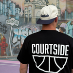 Person wearing a black 'Courtside' t-shirt with a Courtside cap, standing in front of a colourful basketball court mural.