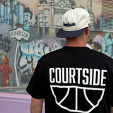 Person wearing a black 'Courtside' t-shirt with a Courtside cap, standing in front of a colourful basketball court mural.