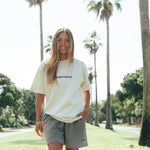 A person wearing a faded bone colored t-shirt and gray shorts standing in a park with palm trees in the background.