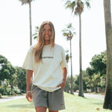 A person wearing a faded bone colored t-shirt and gray shorts standing in a park with palm trees in the background.