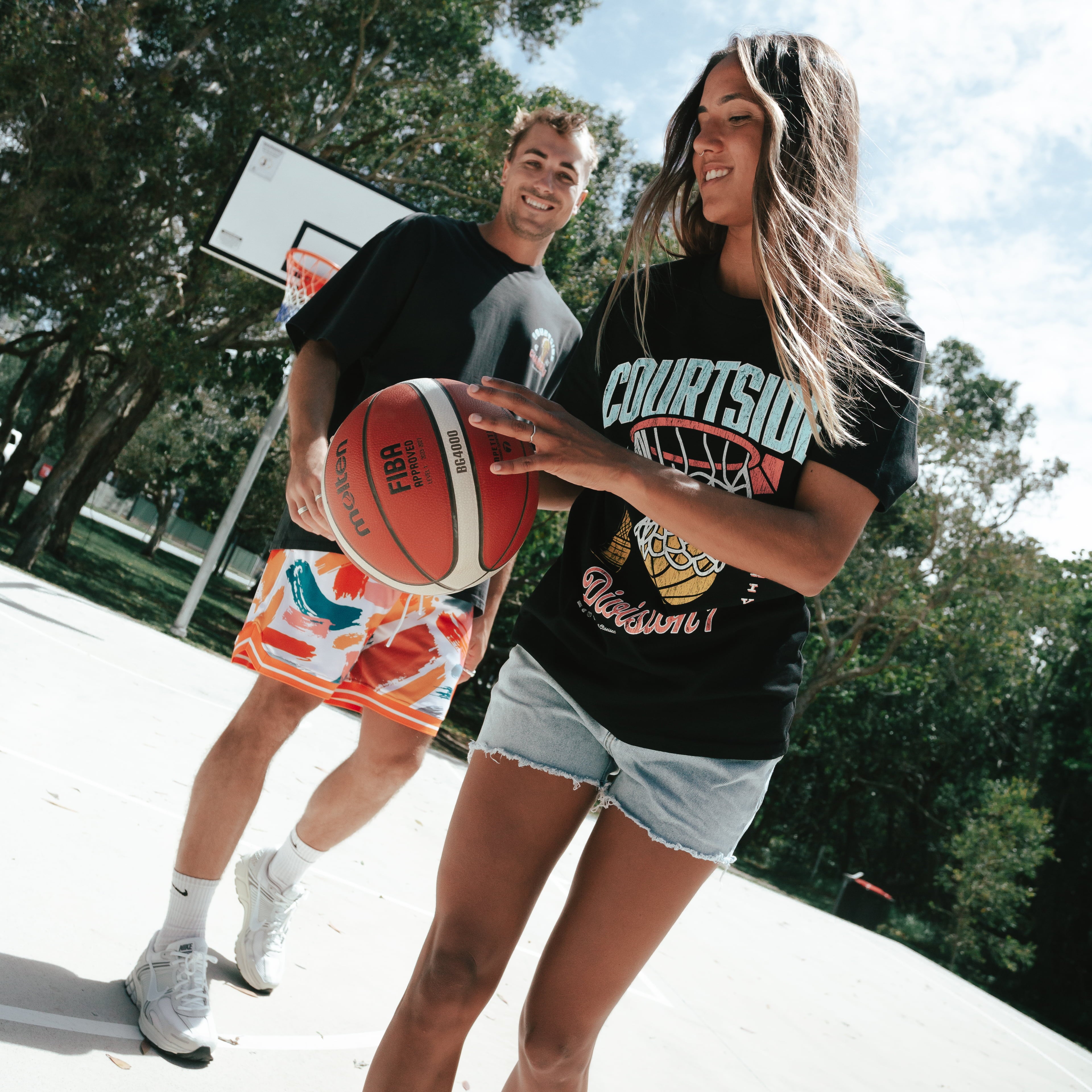 A person wearing a black t-shirt with vintage basketball graphics holding a red and white basketball with man in background on outdoor basketball court