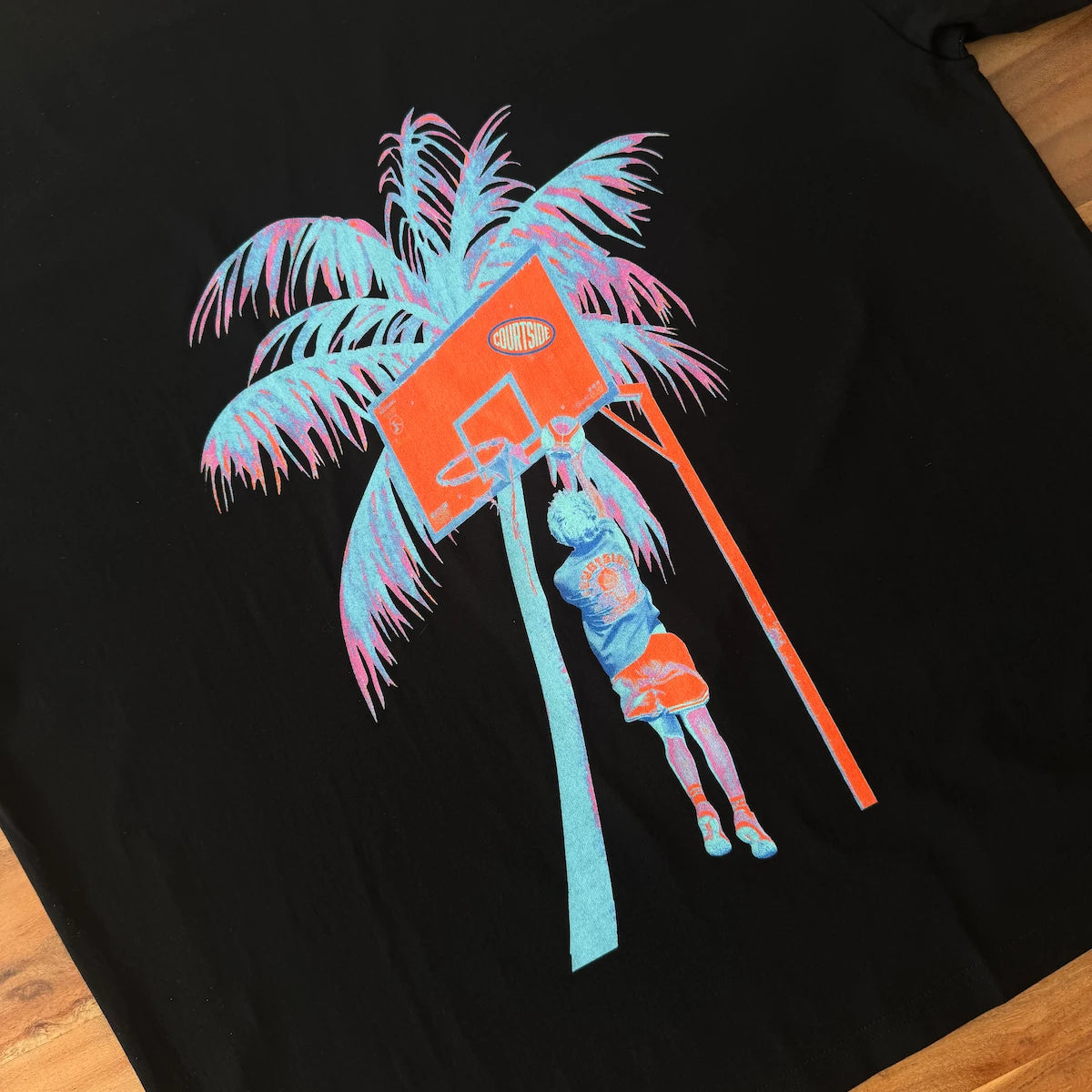 Black tshirt with a colorful palm tree and basketball hoop design on a wooden surface.