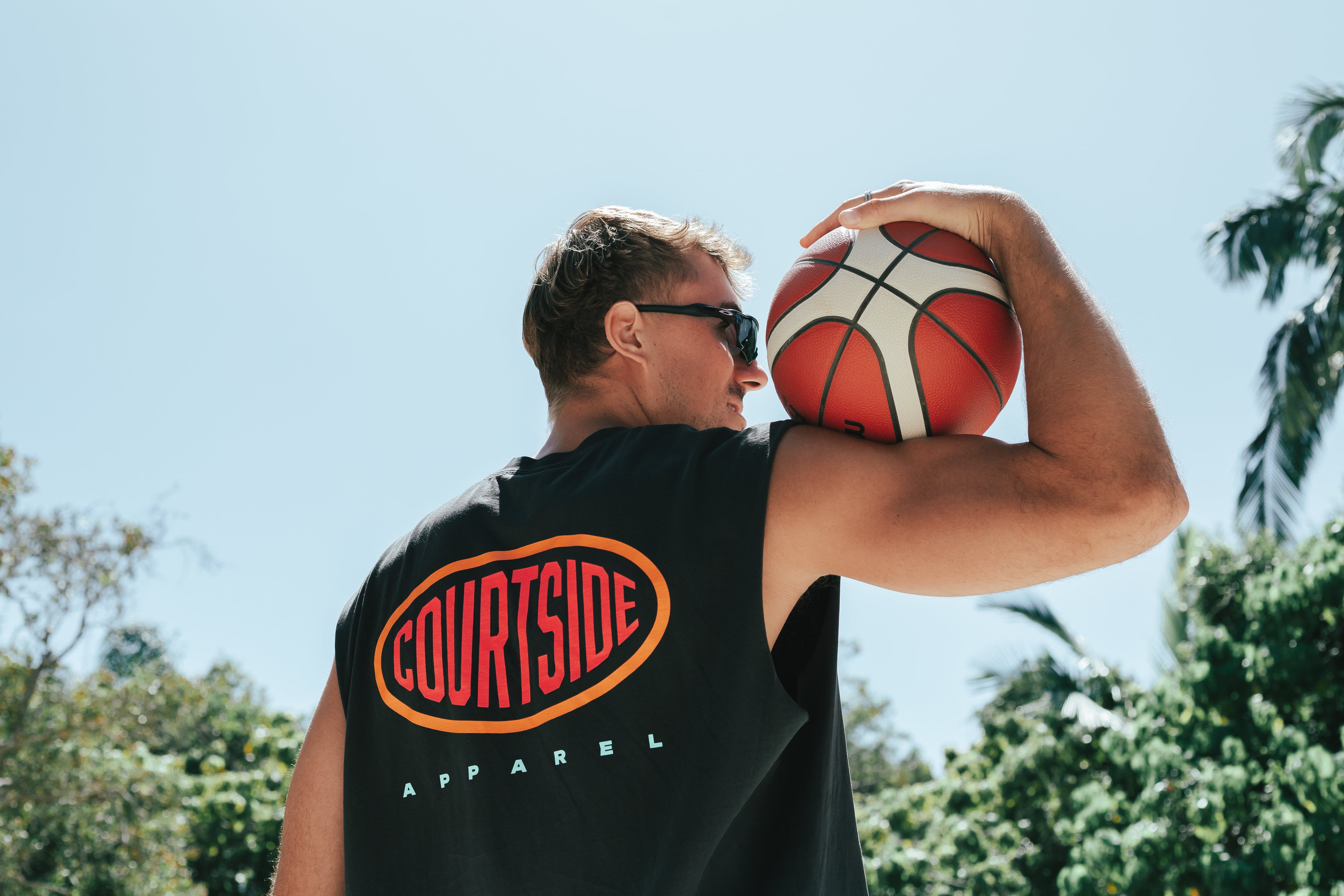 A person wearing sunglasses and a black tank top with the text 'COURTSIDE APPAREL' on the back, holding a red and white basketball.