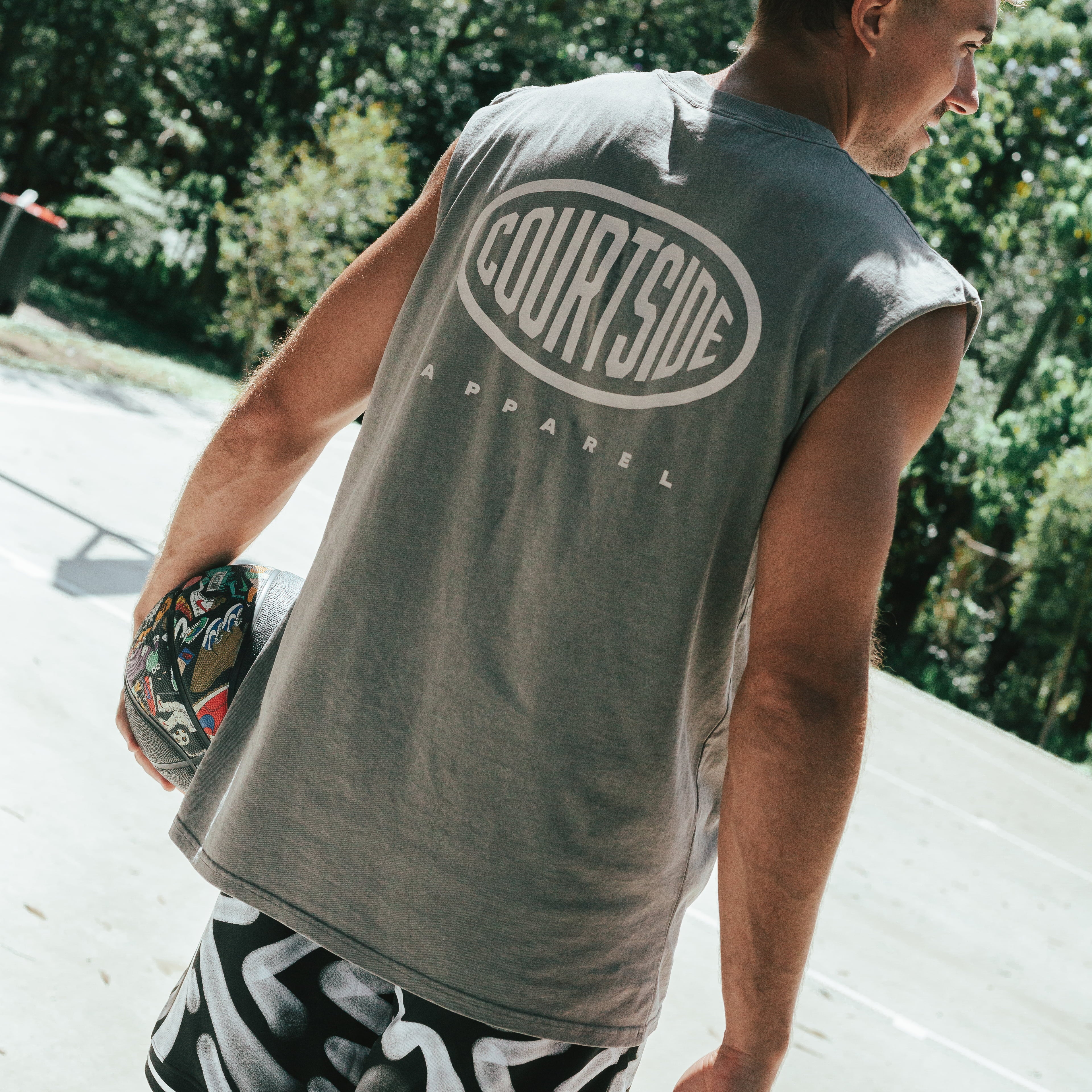 man standing on a basketball court wearing a grey muscle tank and black & white basketball shorts holding a basketball on an outdoor court