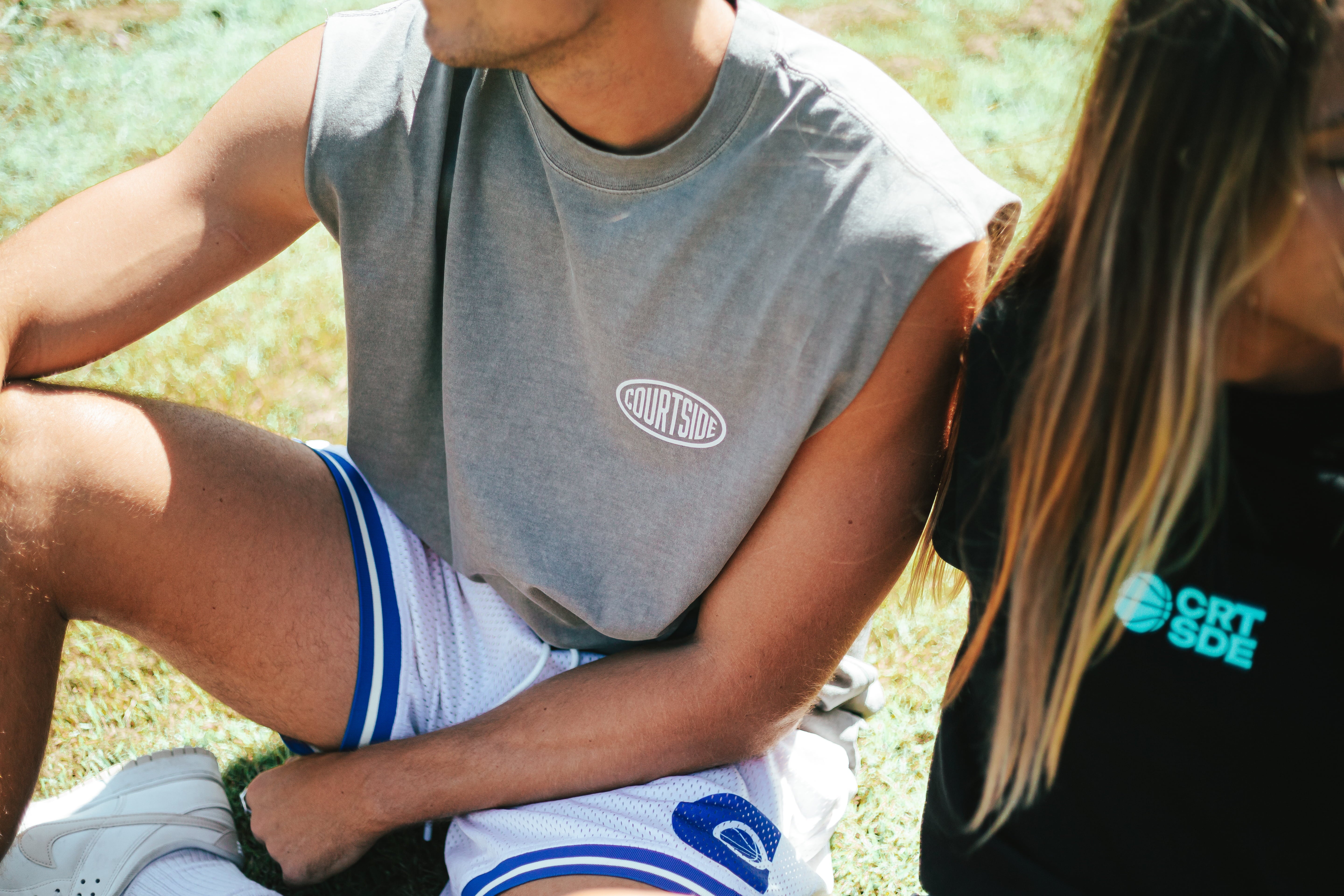 man sitting on ground with grey tank top and cream basketball shorts sitting next to a girl wearing a black t-shirt