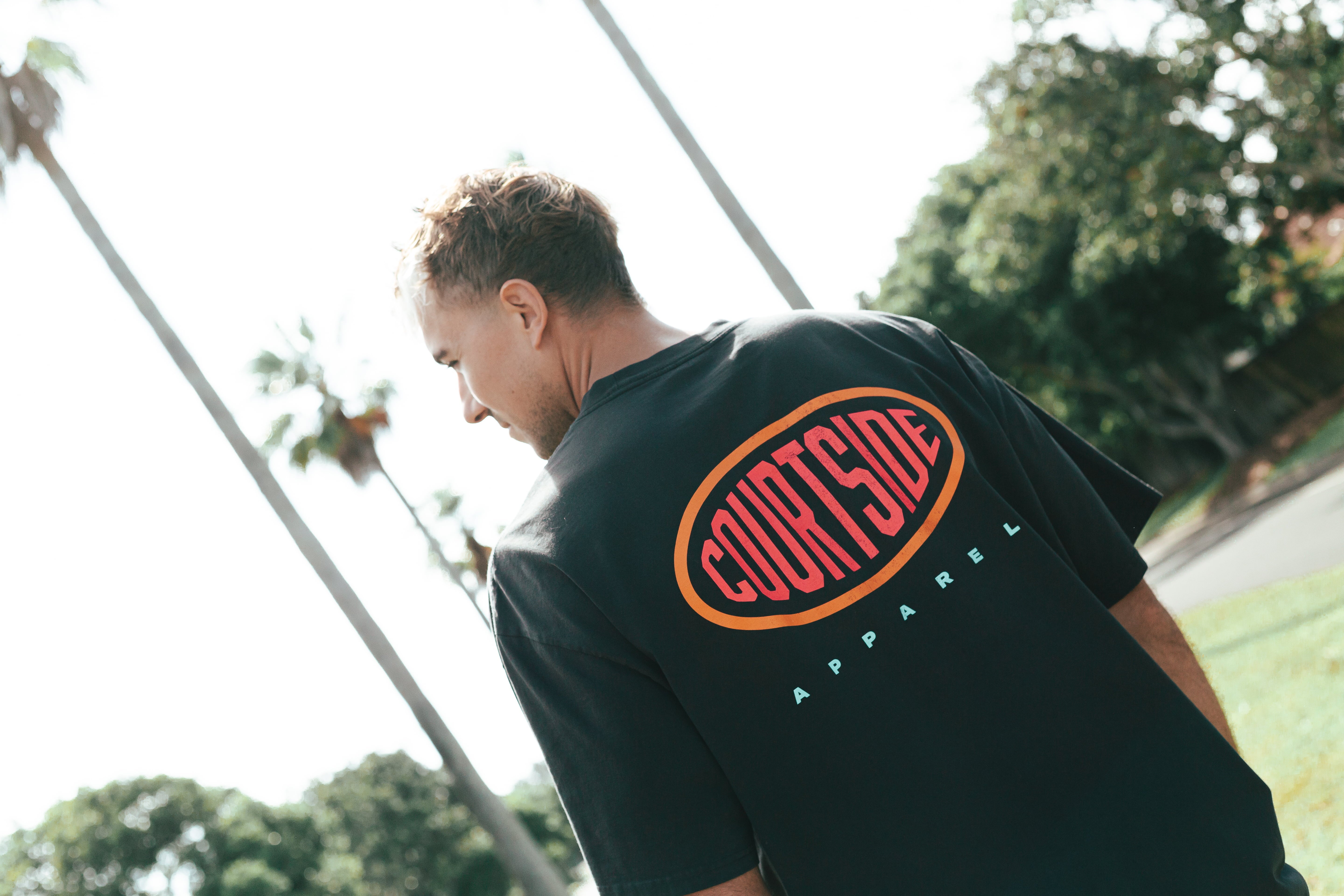 A person wearing a black crew neck T-shirt with the brand logo 'Courtside' on the back.