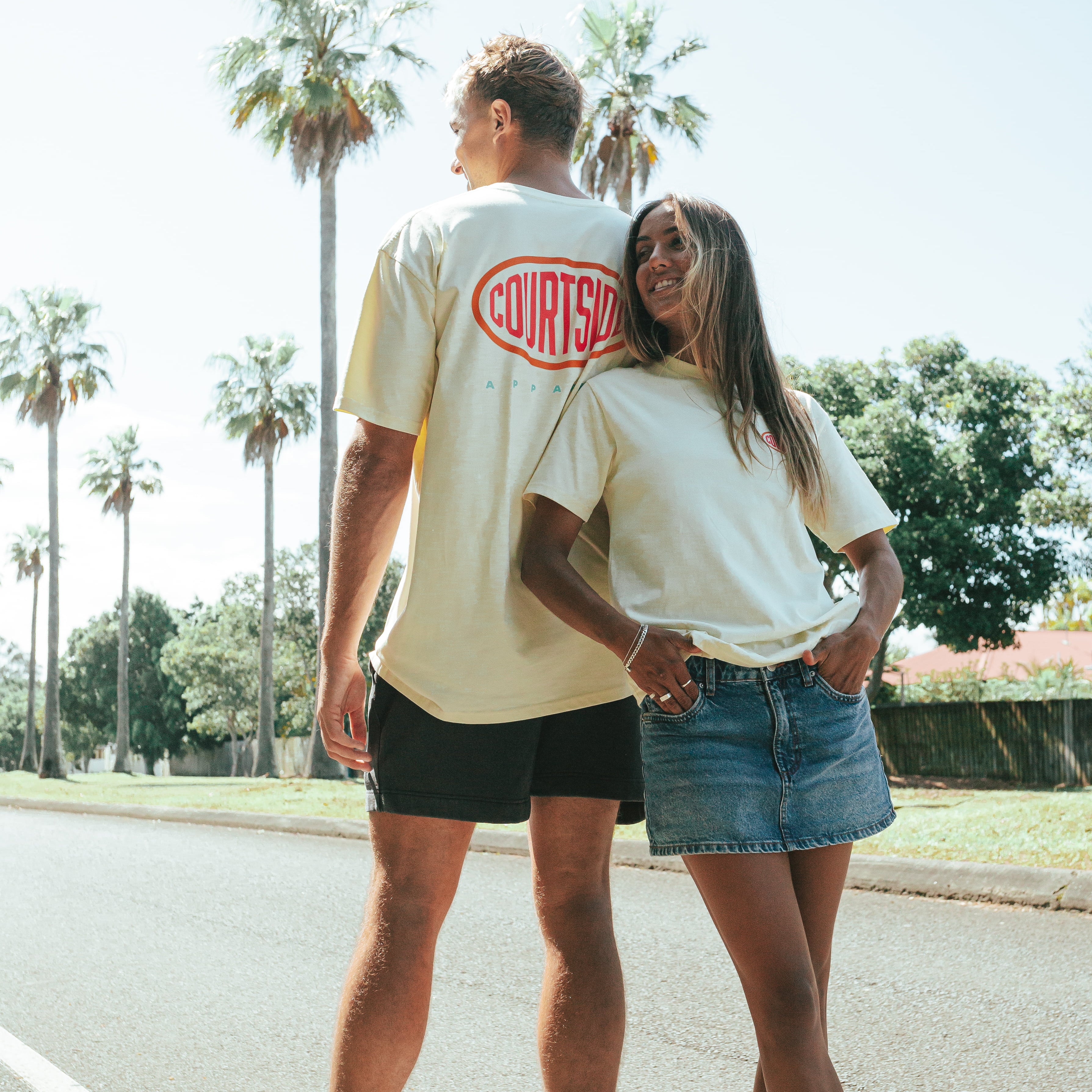 A male and female model wearing matching beige 'Streetball Tee' shirts with a logo on the front, standing on a street with palm trees in the background.