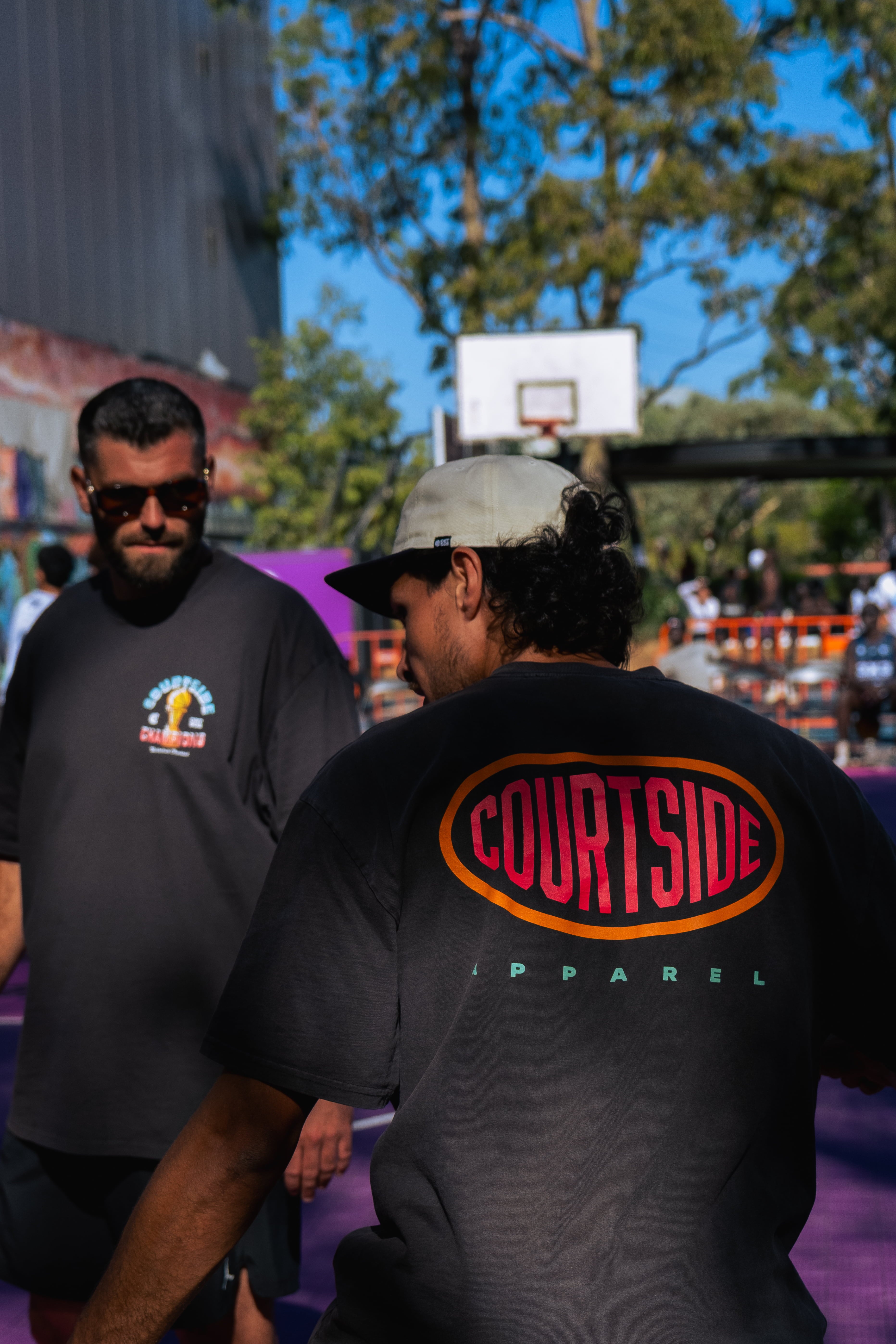 Two men standing on a basketball court in Melbourne with one wearing a 'Courtside Apparel' shirt.