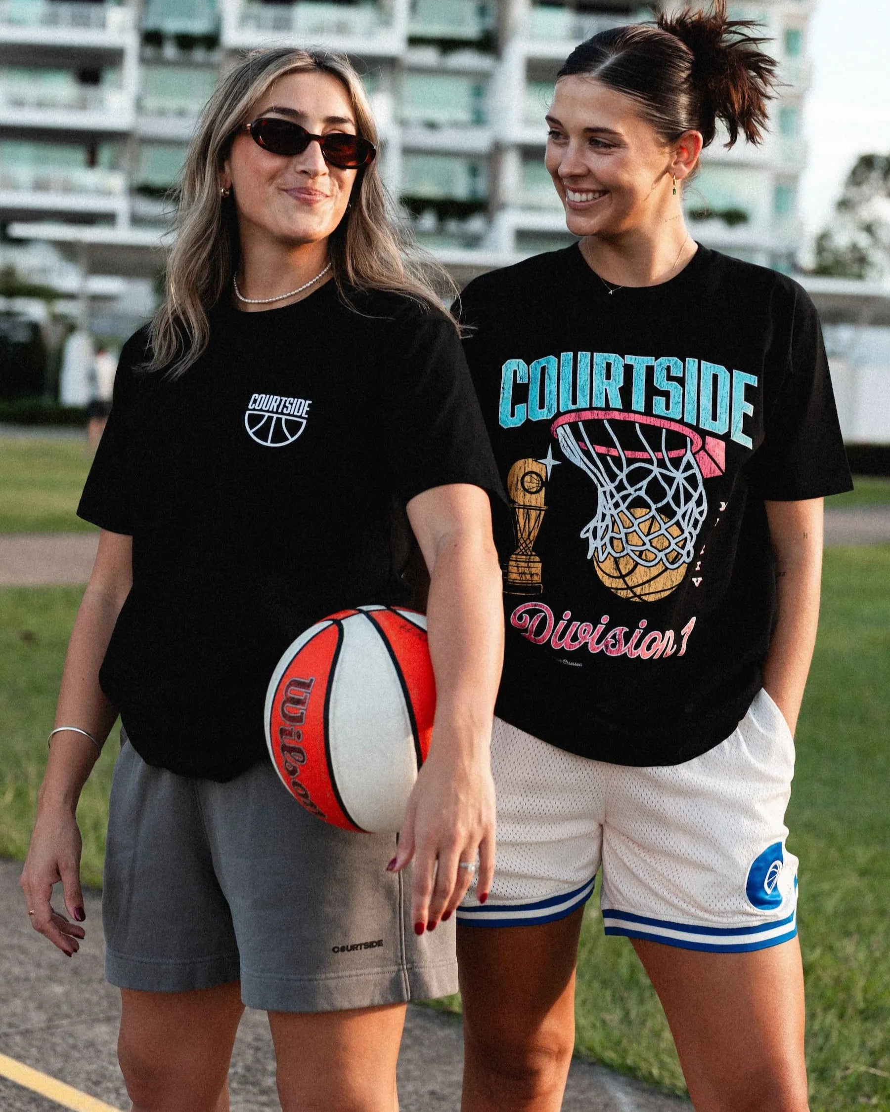 Two women standing outdoors, one holding a basketball, wearing black t-shirts with sports-themed designs.